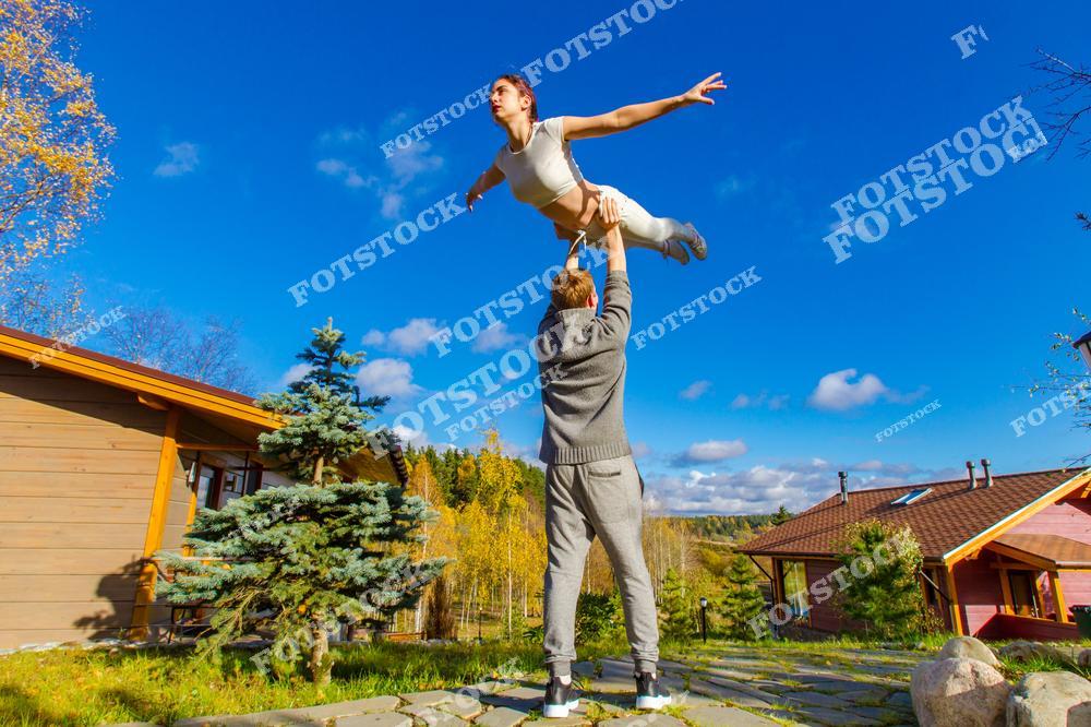 На фотографии мужчина держит девушку высоко над головой на фоне загородного дома и синего неба. Яркий солнечный день и золотая осень создают атмосферу свободы и радости. Снимок символизирует физическую активность, романтику и наслаждение природой.